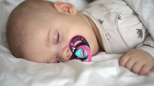 infancy, childhood, development, medicine and health concept - close-up face of newborn chubby sleeping baby 6 month with pacifier lying on stomach covered with blanket on bed on white background.