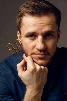 Handsome doctor with surgical scissors close-up. Confident man holding medical equipment in hands and smiling. Happy male posing in studio