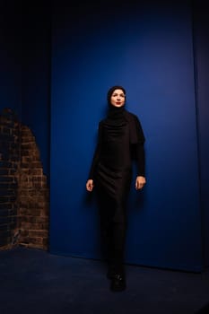 Fashion muslim woman in black hijab is posing on blue background in studio. Professional caucasian model