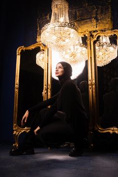 Muslim girl in black hijab near mirrors with her reflections. Fashion muslim model near big expensive chandelier. Islamic religion