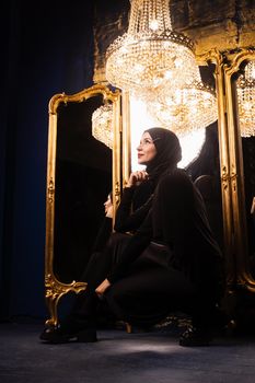 Muslim girl in black hijab near mirrors with her reflections. Fashion muslim model near big expensive chandelier. Islamic religion