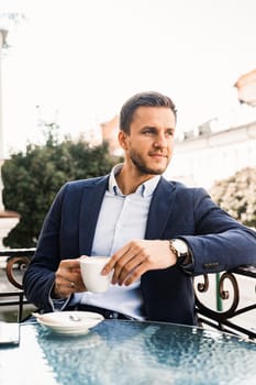 Man is sitting on the summer terrace in cafe, drinking coffee and smiling. Handsome man with cup of coffee in cafe. Morning lifestyle of male