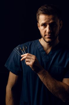 Handsome doctor with surgical scissors on dark background. Confident man holding medical equipment in hands and smiling. Happy male posing in studio