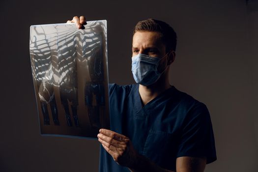Doctor in medical mask for protection of coronavirus covid-19 holding x-ray scan of ill patient. Computered tomography. Surgeon in medical robe looking at picture of scan on dark background.