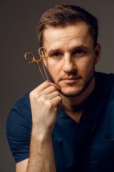 Handsome doctor with surgical scissors close-up. Confident man holding medical equipment in hands and smiling. Happy male posing in studio