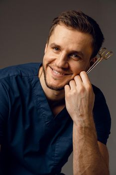 Handsome doctor with surgical scissors close-up. Confident man holding medical equipment in hands and smiling. Happy male posing in studio