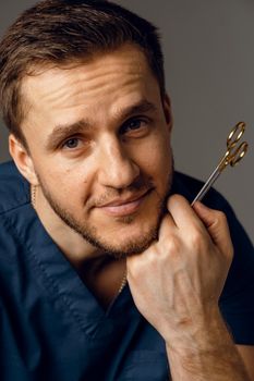 Handsome doctor with surgical scissors close-up. Confident man holding medical equipment in hands and smiling. Happy male posing in studio