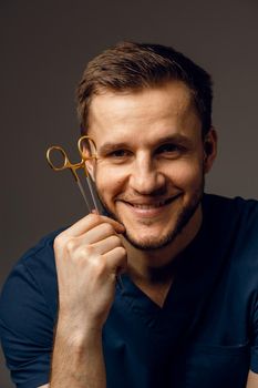 Handsome doctor with surgical scissors close-up. Confident man holding medical equipment in hands and smiling. Happy male posing in studio