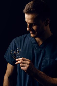 Handsome doctor with surgical scissors on dark background. Confident man holding medical equipment in hands and smiling. Happy male posing in studio