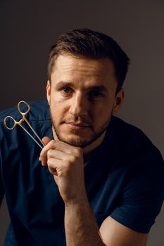 Handsome doctor with surgical scissors close-up. Confident man holding medical equipment in hands and smiling. Happy male posing in studio