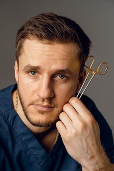 Handsome doctor with surgical scissors close-up. Confident man holding medical equipment in hands and smiling. Happy male posing in studio