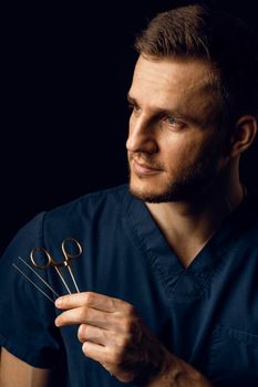 Handsome doctor with surgical scissors on dark background. Confident man holding medical equipment in hands and smiling. Happy male posing in studio