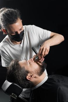 Mustache shaving in barbershop. Barber with dreadlocks in black medical mask trim beard of handsome man at quarantine coronavirus covid-19