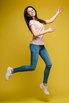 Full length isolate portrait of positive brunette young girl in jeans and t-shirt and sneakers jumping over yellow background. She is smiling at camera.