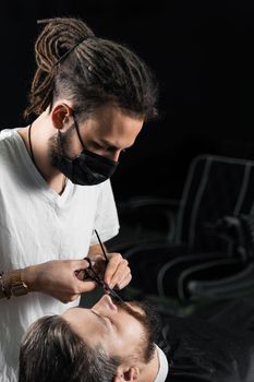 Mustache shaving in barbershop. Barber with dreadlocks in black medical mask trim beard of handsome man at quarantine coronavirus covid-19