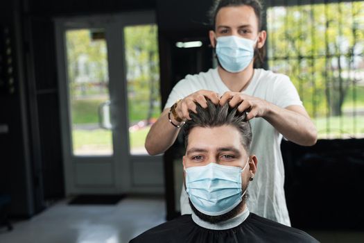 Hair styling with wax for a bearded man in a medical mask. The work of the barbershop in quarantine coronavirus covid-19.