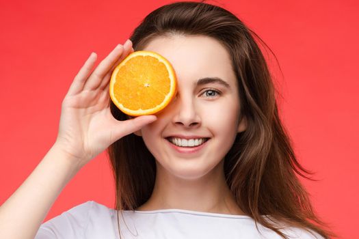 Front view of cheerful young blonde posing with fresh oranges on isolated background. Funny girl keeping fruit and closing eyes in studio. Concept of happiness and health.