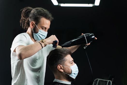 Barber in medical mask dries the hair of a handsome bearded man after a fashionable haircut. The work of a hairdresser during the quarantine period coronavirus covid-19