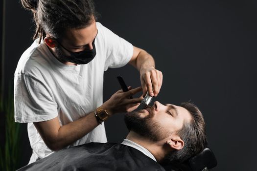 Mustache shaving in barbershop. Barber with dreadlocks in black medical mask trim beard of handsome man at quarantine coronavirus covid-19