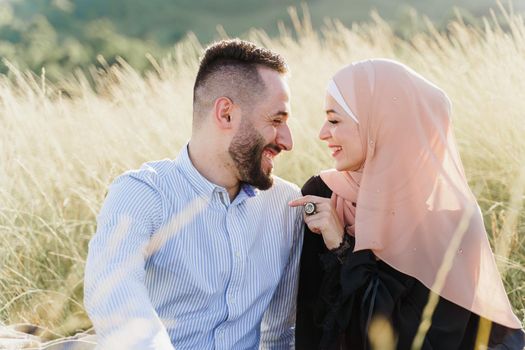 Muslim love story. Mixed couple seats on the grass, smiles and hugs.. Woman weared in hijab looks to her man. Advert for on-line dating agency