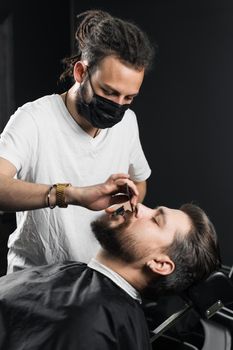 Mustache shaving in barbershop. Barber with dreadlocks in black medical mask trim beard of handsome man at quarantine coronavirus covid-19