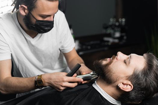 Beard shaving in barbershop. Barber with dreadlocks in black medical mask trim bearded man at quarantine coronavirus covid-19