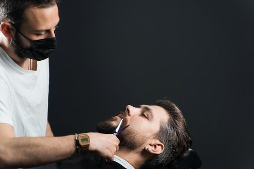 Beard shaving in barbershop. Barber with dreadlocks in black medical mask trim bearded man at quarantine coronavirus covid-19