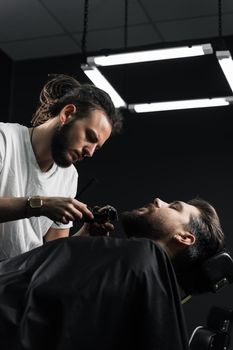 Handsome bearded man has hair and beard cut in barbershop. Dreadlock barber cuts client's hair.