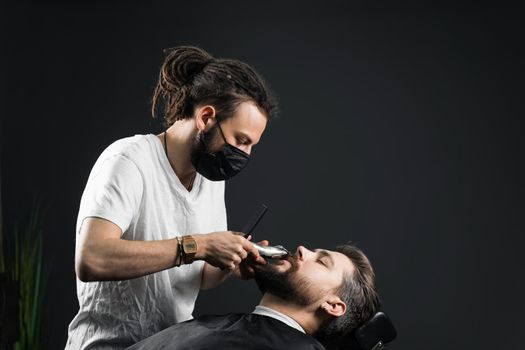 Mustache shaving in barbershop. Barber with dreadlocks in black medical mask trim beard of handsome man at quarantine coronavirus covid-19