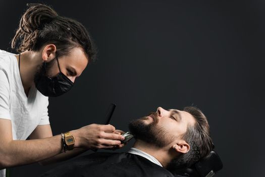 Beard shaving in barbershop. Barber with dreadlocks in black medical mask trim bearded man at quarantine coronavirus covid-19