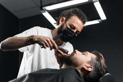 Mustache shaving in barbershop. Barber with dreadlocks in black medical mask trim beard of handsome man at quarantine coronavirus covid-19