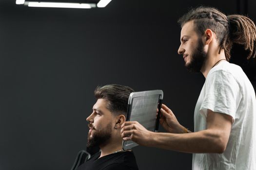 Barber with dreadlocks showing result of low fade machine hair and beard cut for bearded man in barbershop. Hairstyle with a smooth transition