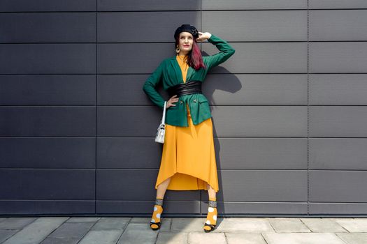 Young woman in bright clothes, yellow skirt and green jacket. Yellow socks in sandals, beret on the head, hair with the color of magenta. Caucasian female fashion model standing against gray wall background, open with copy space