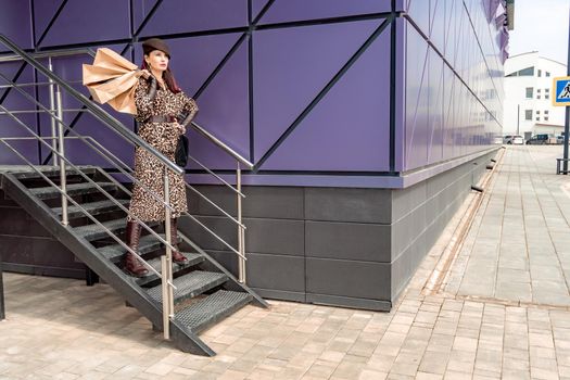 A happy shopaholic girl keeps her bags near the shopping center. A woman near the store is happy with her purchases, holding bags. Dressed in a leopard print dress. Consumer concept