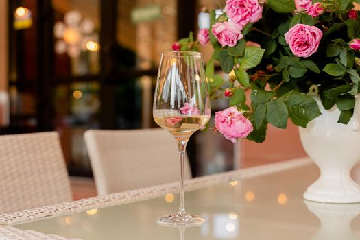 Glass of white wine against the background of bouquet roses in white vase on light table