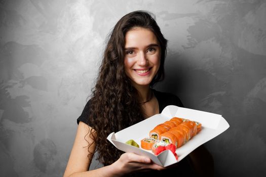 Girl holding philadelphia rolls in a paper box on gray background. Sushi, food delivery