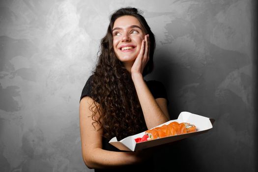 Surprised girl with sushi set philadelphia rolls in a paper box happy girl holding on a gray background. Food delivery.