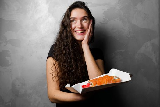 Surprised girl with sushi set philadelphia rolls in a paper box happy girl holding on a gray background. Food delivery