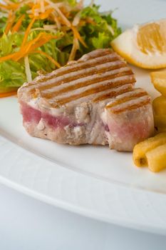 Tuna steak grilled with french fries salad of carrots, parsnips, lettuce and lemon on white plate on white background