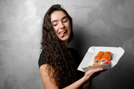 Surprised girl with sushi set philadelphia rolls in a paper box happy girl holding on a gray background. Food delivery