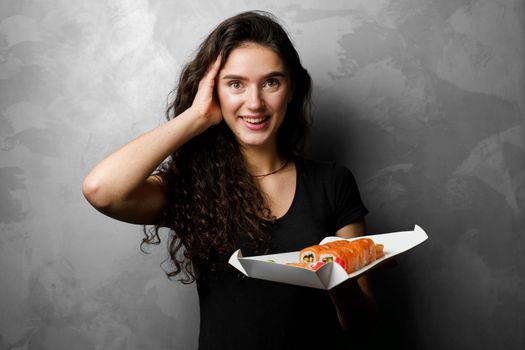 Surprised girl with sushi set philadelphia rolls in a paper box happy girl holding on a gray background. Food delivery