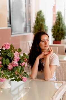 Dreaming woman with glass of alcoholic white wine in cafe in summer terrace and bouquet of roses on the table.