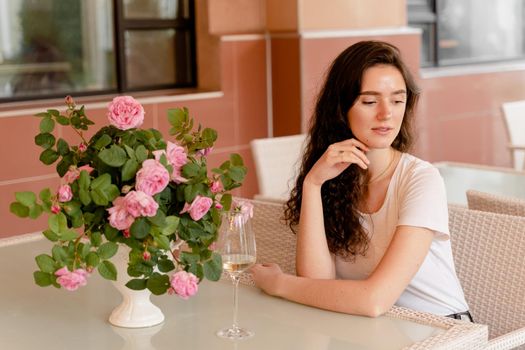 Dreaming woman with glass of alcoholic white wine in cafe in summer terrace and bouquet of roses on the table.