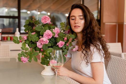 Dreaming woman with glass of alcoholic white wine in cafe in summer terrace and bouquet of roses on the table