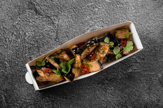 Fried eggplant dish in cardboard box. Food delivery. Vegetables garnished with cilantro and chili on gray background