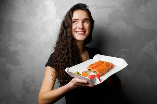 Girl holding philadelphia rolls in a paper box on gray background. Sushi, food delivery