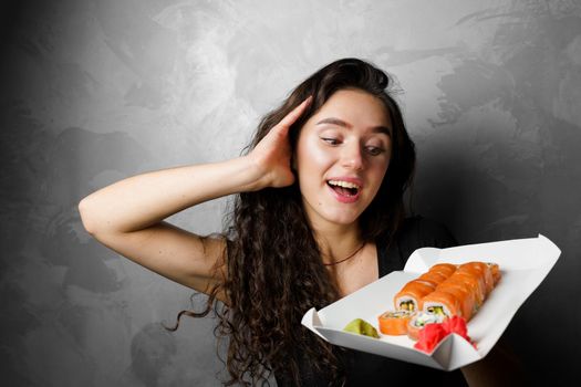 Surprised girl with sushi set philadelphia rolls in a paper box happy girl holding on a gray background. Food delivery