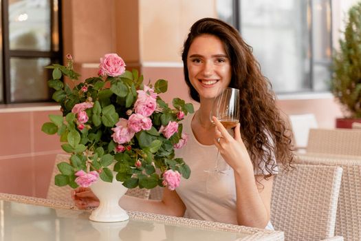 Dreaming woman with glass of alcoholic white wine in cafe in summer terrace and bouquet of roses on the table