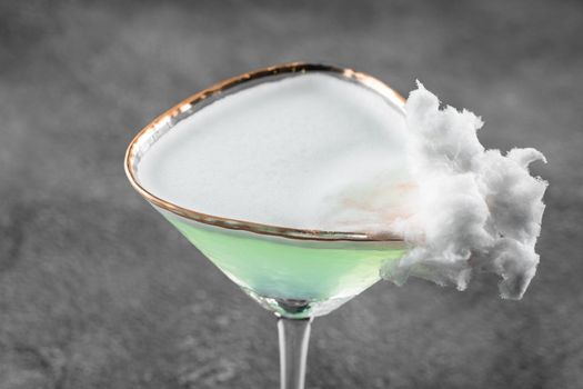 Green alcohol cocktail in a glass decorated with cotton candy on gray background