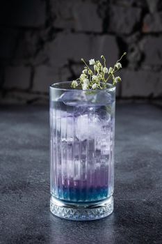 A cocktail in a tall glass with blue liqueur and ice, decorated with white flowers on gray background